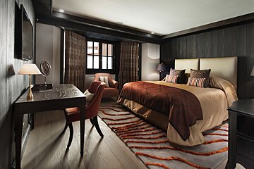 A stylish bedroom with dark wood walls, a large bed, orange accent chair, patterned rug, and a desk area by a window.