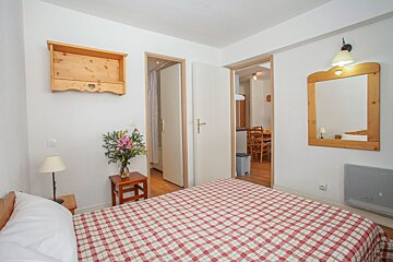 A cozy bedroom features a red checkered bedspread, wooden mirror and shelf, and flowers. Open doorways lead to a bathroom and kitchen/dining area.