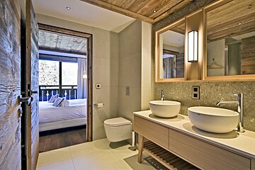 A modern bathroom with dual sinks, a toilet, and rustic wood accents. An open door reveals a bedroom with a balcony and mountain views.