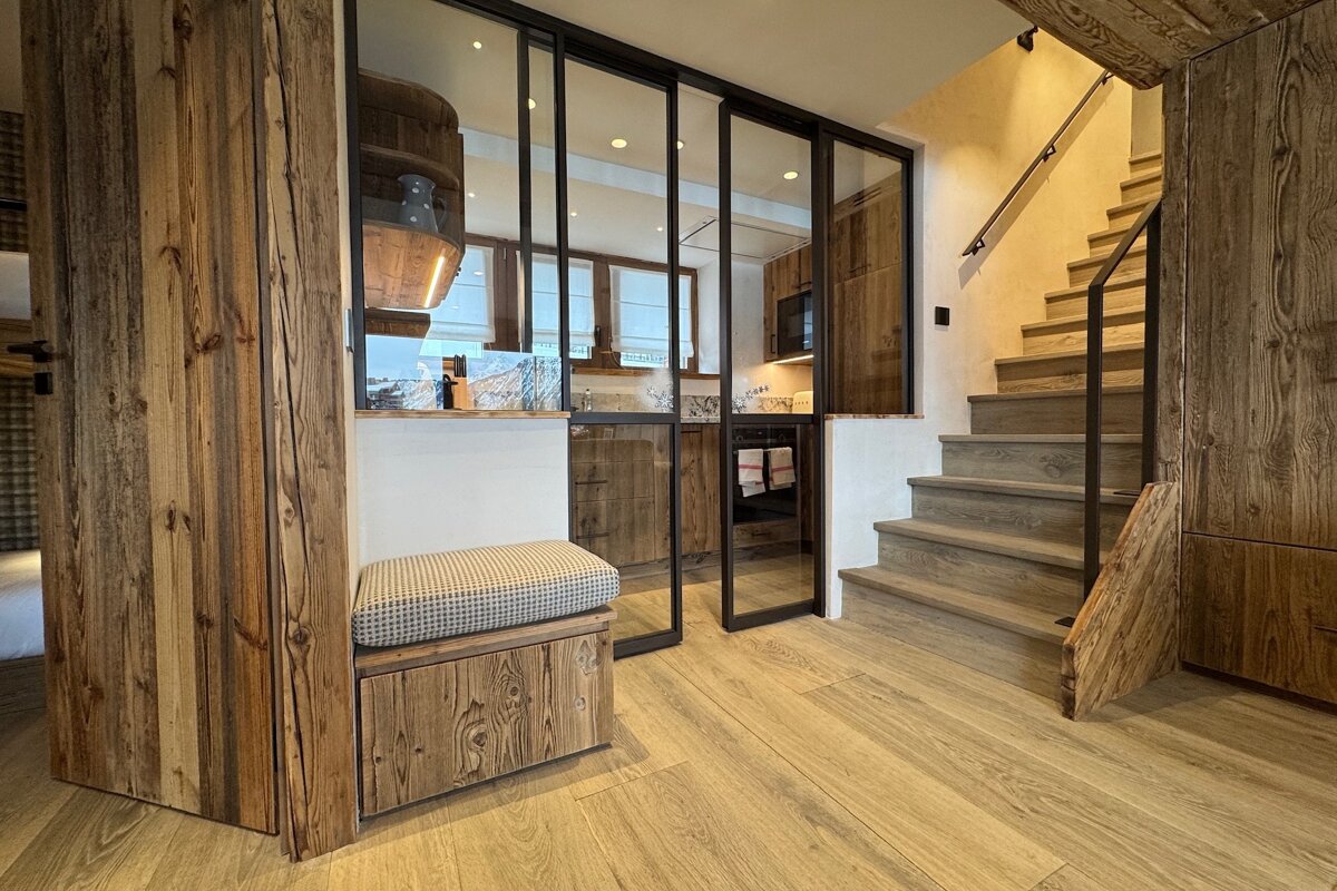 Modern rustic chalet interior featuring a wooden staircase, a small bench, and glass sliding doors looking into a kitchen.