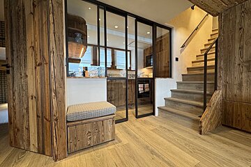Modern rustic chalet interior featuring a wooden staircase, a small bench, and glass sliding doors looking into a kitchen.