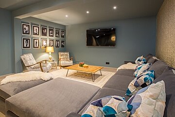 Modern living room with a large grey sectional, blue walls, a wall-mounted TV, two armchairs, and a gallery wall of black-and-white photos.