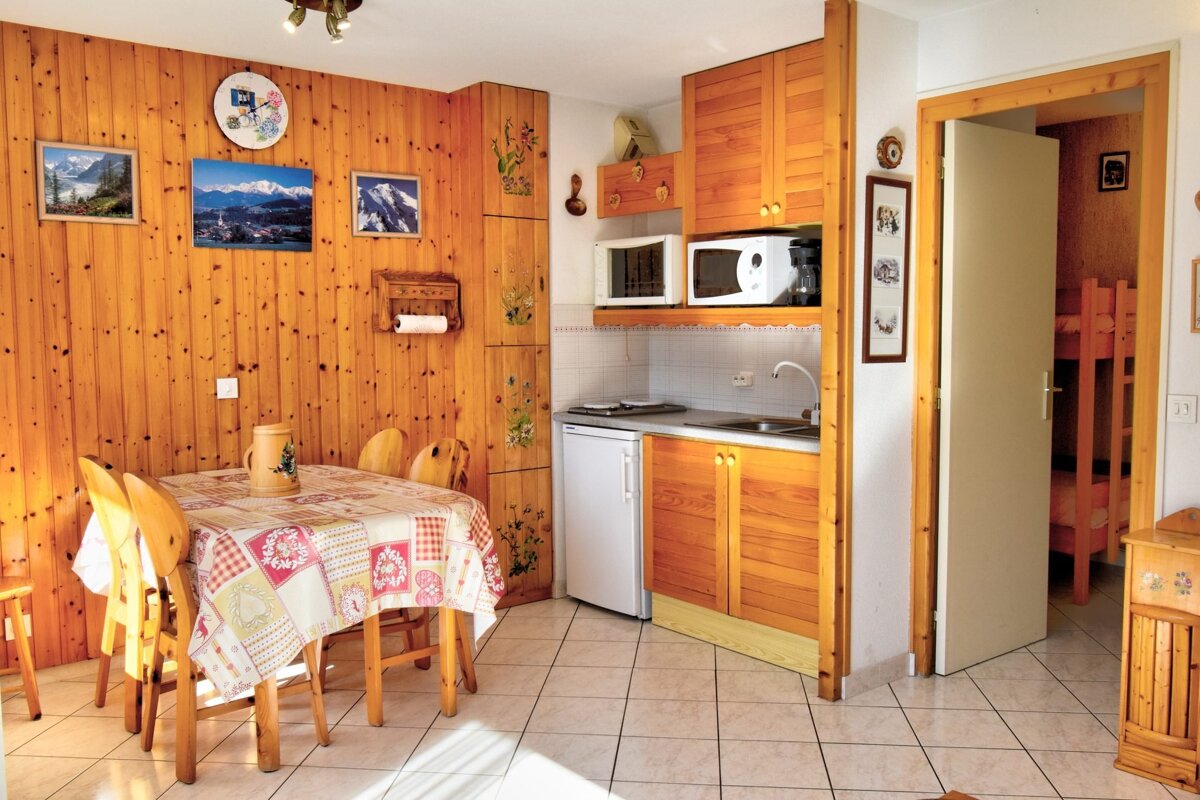 A cozy, wood-paneled kitchenette and dining area with mountain pictures, leading to a room with bunk beds. Features a small fridge, microwave, and patterned tablecloth.