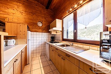 A cozy, rustic wooden kitchen with white tiled backsplash, a window offering mountain views, and essential appliances like a fridge, stove, and kettle.