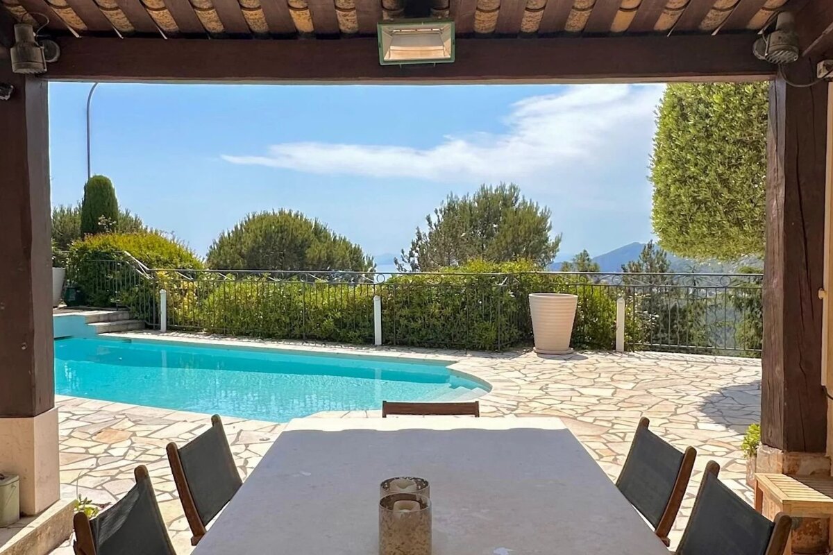 Covered patio with a dining table looking out at a bright blue swimming pool, stone terrace, and scenic hills.