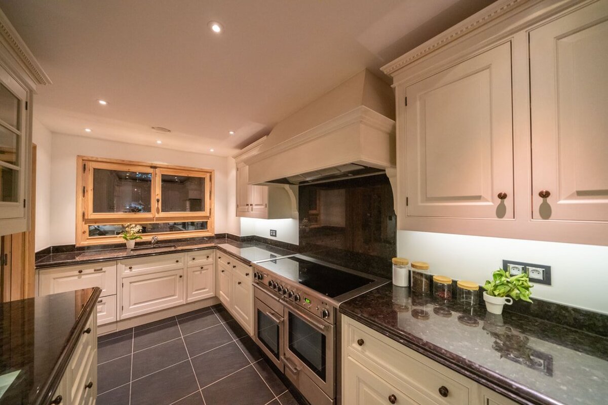 A luxurious kitchen featuring cream cabinets, dark granite countertops, a large stainless steel range, dark tiled floors, and wooden window frames.