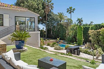 Sunny house with a balcony overlooking a lush, terraced garden featuring a swimming pool, palm trees, and various outdoor seating areas.