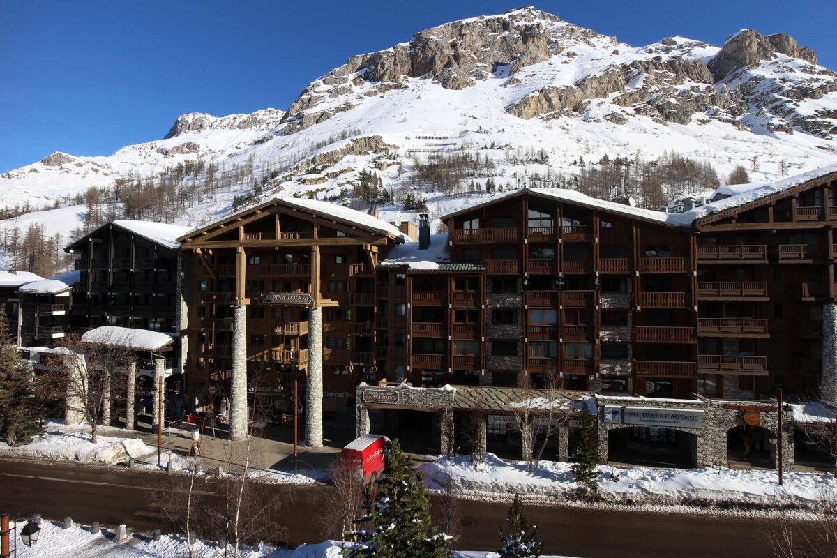 Rustic wooden ski resort buildings stand against majestic snow-covered mountains under a clear blue sky, creating a beautiful winter scene.