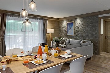 A modern open-plan living and dining room with a full breakfast spread on the wooden table and a cozy grey sofa in the background.
