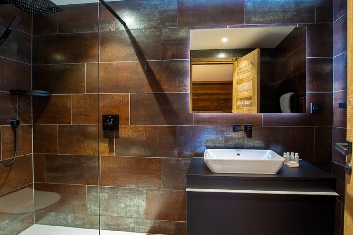 Modern bathroom with dark rust-colored tiles, a glass shower, and a black vanity with a white vessel sink under a backlit mirror.