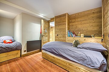 A cozy, chalet-style bedroom features two single beds, rustic wood-paneled walls with built-in shelves, and a vibrant red striped floor.