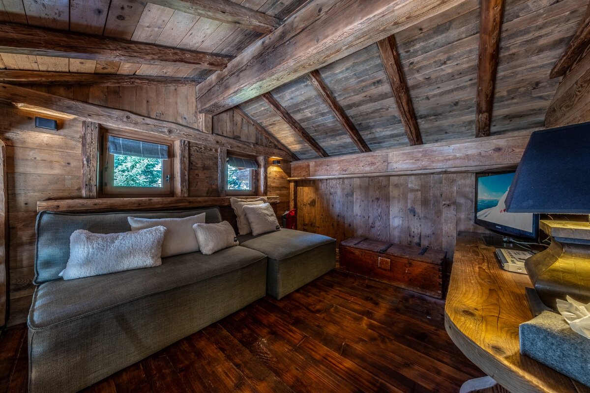 Cozy, rustic cabin-style room with natural wood walls, ceiling, and floor. Features a plush sofa, windows, and a TV with a lamp.