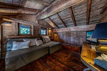 A cozy, rustic wooden room with exposed beams, a comfortable sofa, small windows, and a desk with a TV, creating a warm cabin feel.
