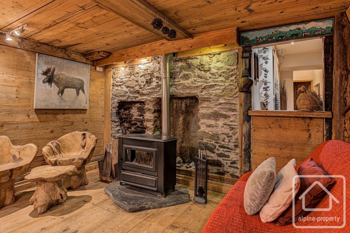 A cozy, rustic chalet living room with wood paneled walls, a stone fireplace, wood stove, moose art, carved wooden furniture, and a red sofa.