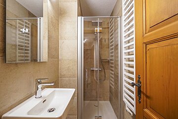 A modern, compact bathroom featuring beige tiles, a white ceramic sink, a glass shower enclosure, and a wooden door.