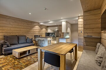 Modern open-plan living room and kitchen with warm wood paneling, gray sofa, wooden dining table, and sleek breakfast bar.