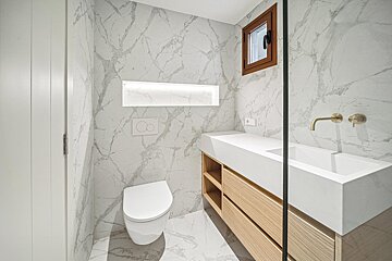 Sleek marble bathroom with a wall-mounted toilet, wooden vanity & sink with brass fixtures, an illuminated niche, and a small window.