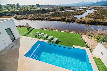 A modern house with a bright blue pool, green lawn, and lounge chairs, overlooking a marshy river landscape with distant mountains.