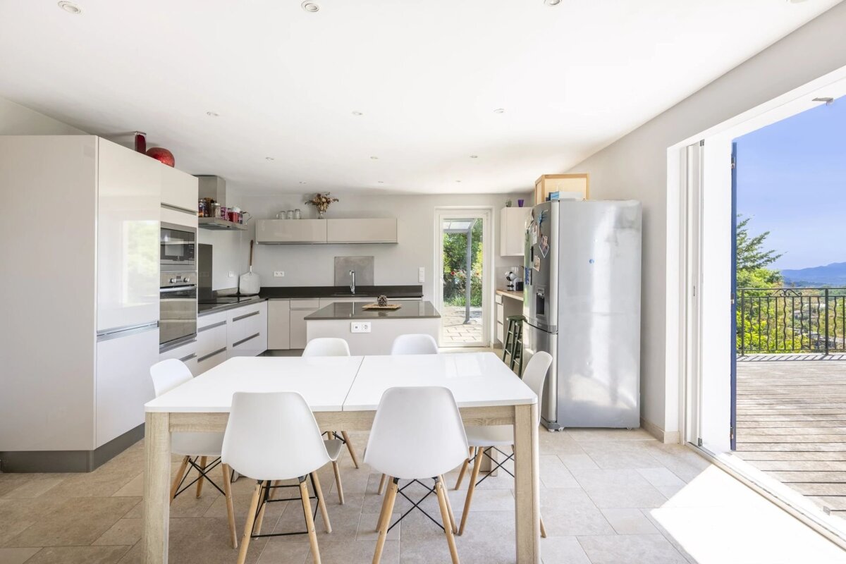 A modern, bright kitchen and dining area with white cabinets, a table with six chairs, and a large open doorway leading to a scenic balcony.