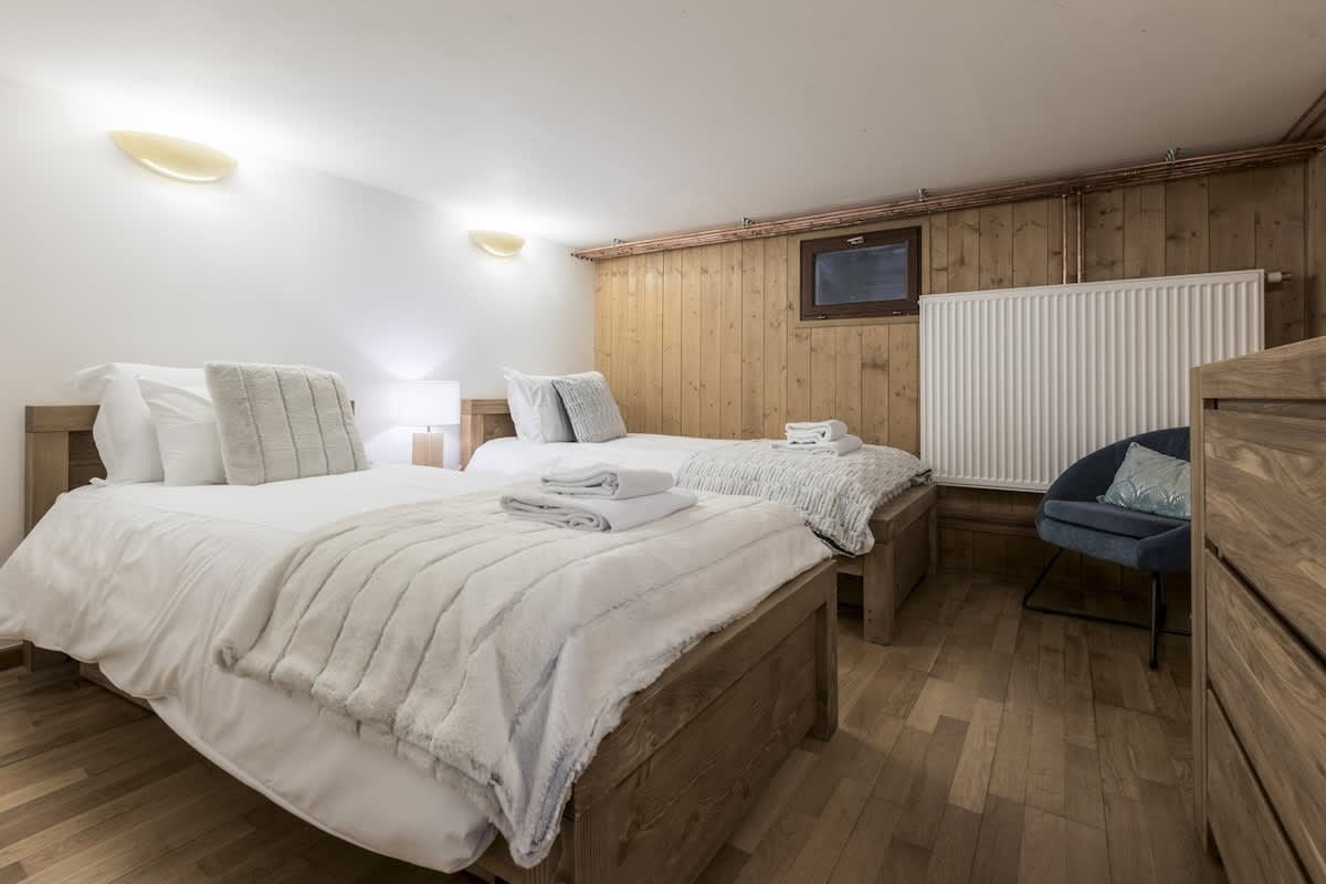 Cozy cabin-style bedroom with two single beds, white bedding, wood-paneled walls, hardwood floors, and a blue accent chair.
