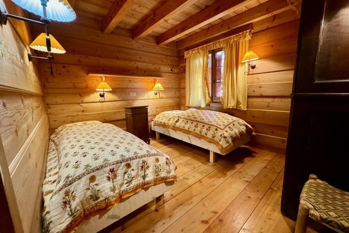 A cozy log cabin bedroom featuring two twin beds with floral quilts, warm sconce lighting, and wooden walls, ceiling, and floors.