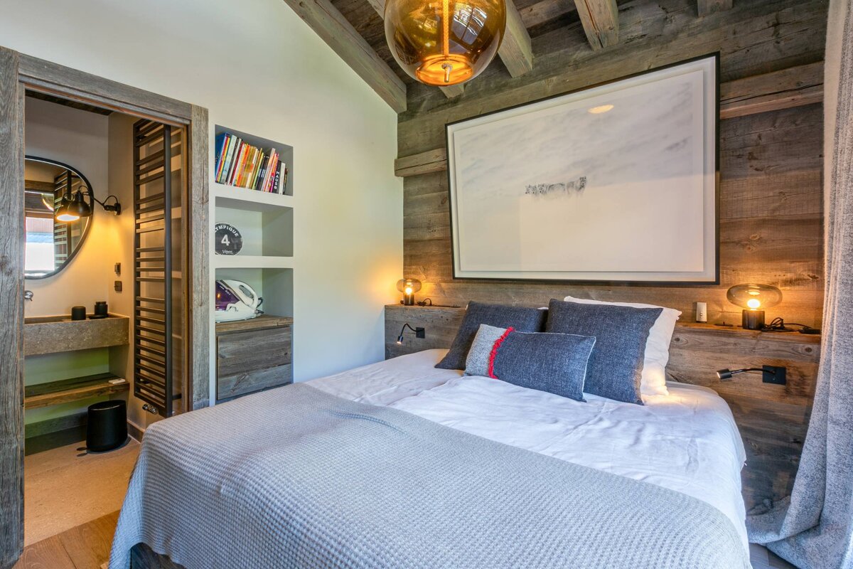 A warm, rustic modern bedroom with rich wood paneling, exposed beams, and a comfortable bed. An en-suite bathroom is visible to the left.