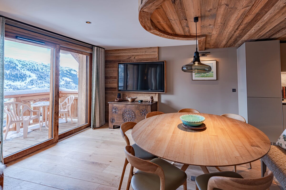Rustic-chic dining room with a round wooden table, wood-paneled walls, and large sliding glass doors showing a snowy mountain view.