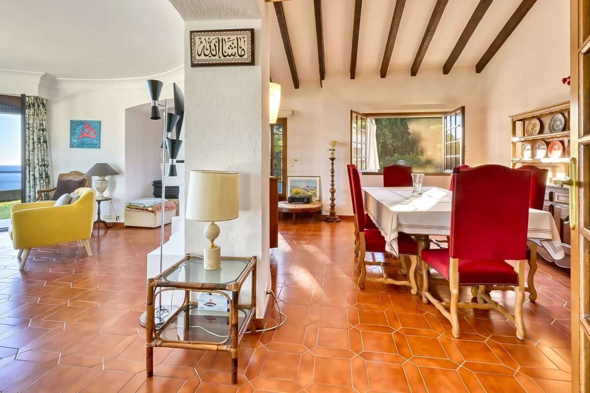 Spacious living & dining room with terracotta floors. Yellow armchair faces sea view. Dining area features red chairs, white table, & wooden beamed ceiling.