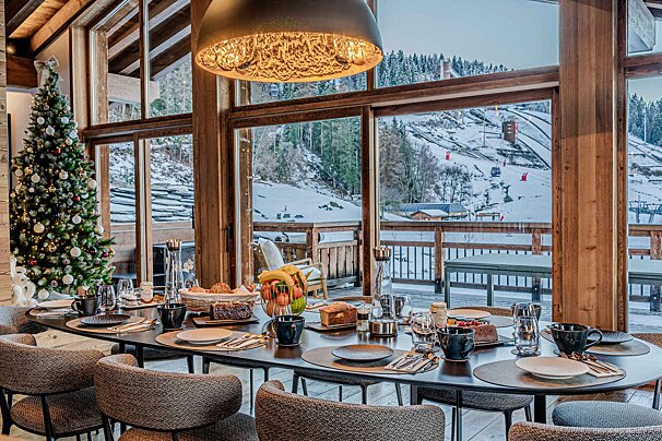 Festive breakfast table in a cozy alpine chalet with floor-to-ceiling windows showing snowy ski slopes and a decorated Christmas tree.