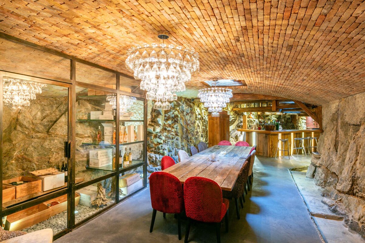A luxurious wine cellar features a brick vaulted ceiling, stone walls, wine glass chandeliers, a long wooden table with red chairs, glass wine storage, and a bar.
