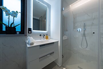 A modern bathroom featuring a white vanity, illuminated mirror, glass shower, and a white orchid by the window.