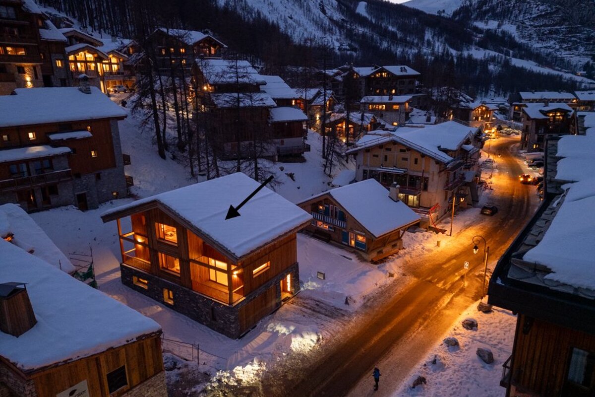 Snowy alpine village at dusk with warmly lit wooden chalets and a black arrow pointing to a snow-covered roof.