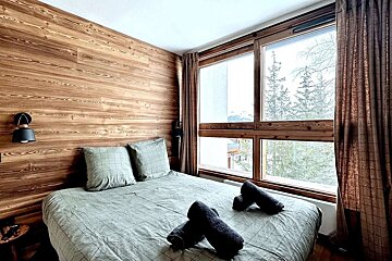A cozy, wood-paneled bedroom with a bed and green bedding. A large window offers a view of snowy pine trees and a distant mountainous landscape.