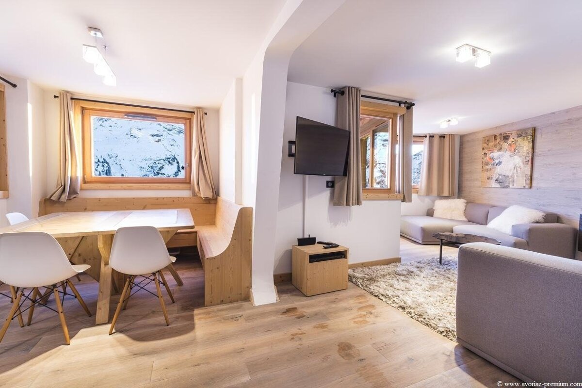 Modern alpine chalet interior featuring a dining nook with mountain views and an adjacent living room with grey sofas and a TV.
