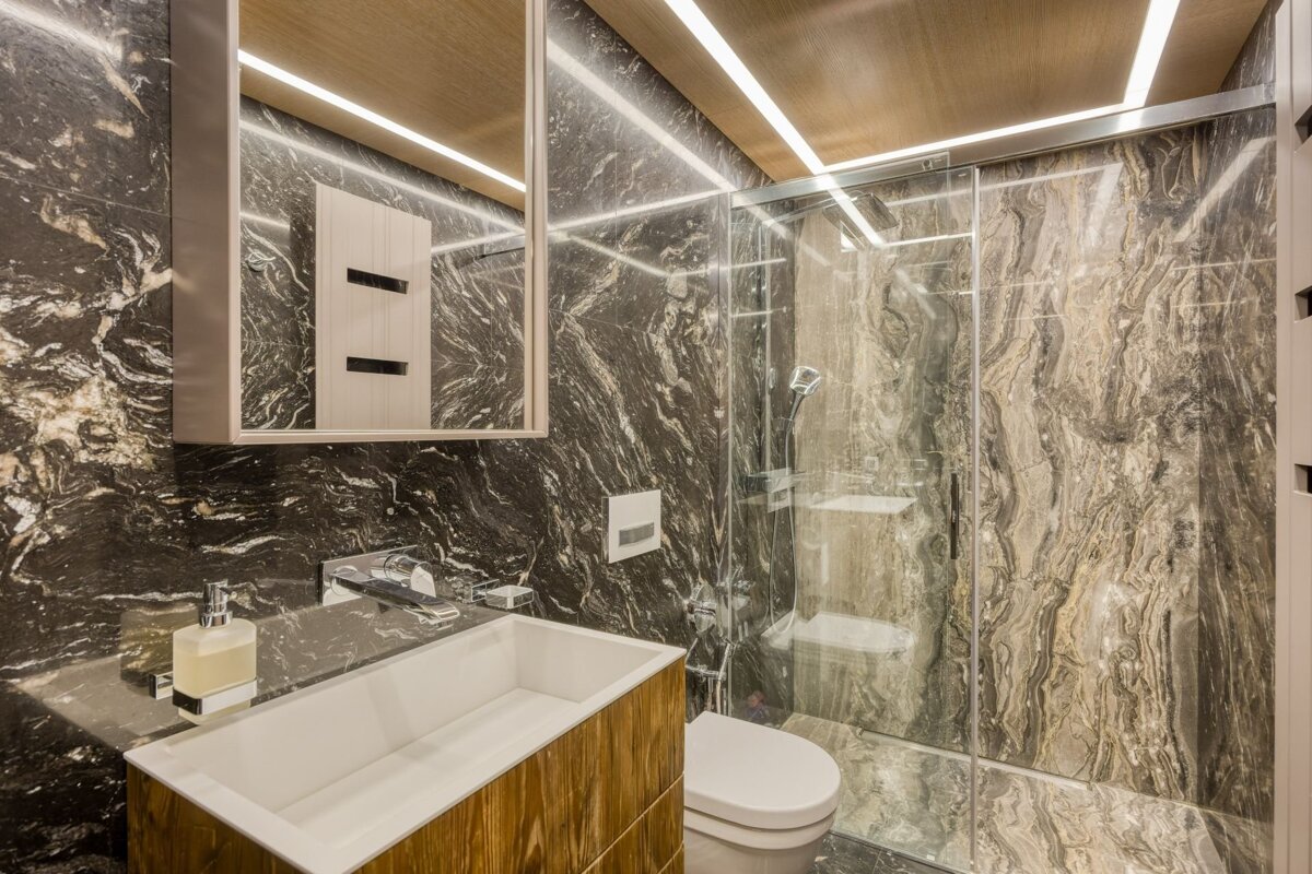 A modern bathroom with dark veined marble walls, wooden ceiling with strip lighting, a glass shower, white sink with wooden vanity, and toilet.