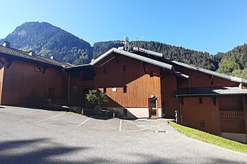 A large wooden chalet-style building with a parking lot, set against a backdrop of forested mountains under a clear blue sky.