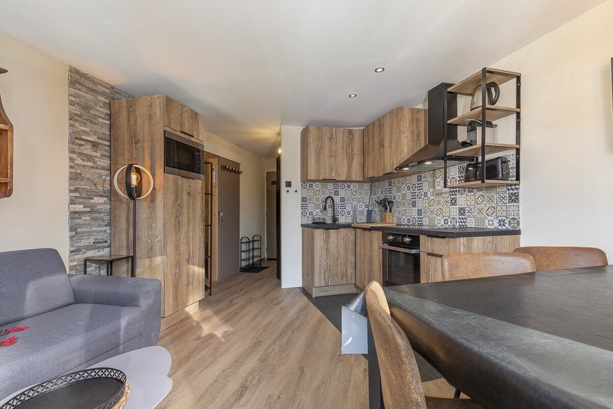 A modern open-plan living space with a kitchen featuring patterned tiles, wooden cabinets, a dining table, and a grey sofa.
