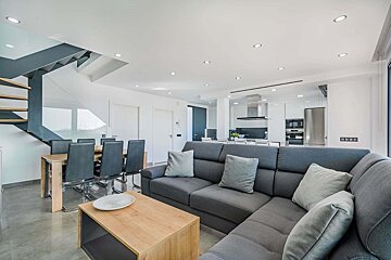 A modern, open-plan living room, dining area, and kitchen with a grey sectional, wooden table, and sleek appliances. A contemporary staircase is on the left.