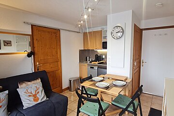 A cozy studio apartment with a kitchenette, dining table set for two, a sofa with deer-themed pillows, and rustic wooden doors.