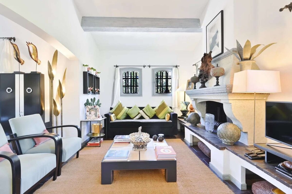 A bright, eclectic living room featuring a large stone fireplace, dark sofa with green cushions, and light blue armchairs. Various artworks and books decorate the space.