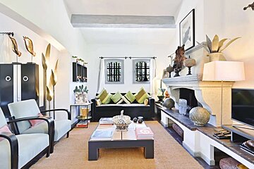 A bright, eclectic living room with white walls, a prominent stone fireplace, a dark sofa with green pillows, light armchairs, and various unique golden and dark decorative elements.