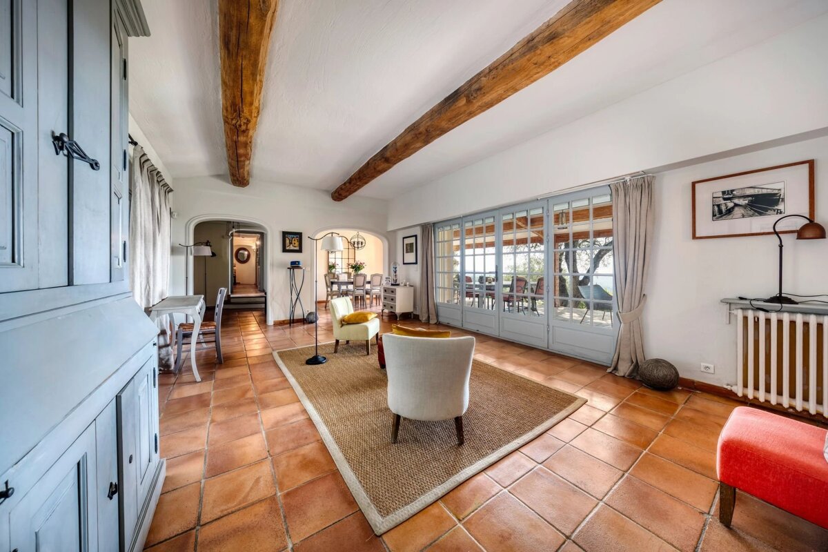 Rustic living room with terracotta tile floors, exposed wood beams, french doors, a neutral rug, and cozy armchairs.