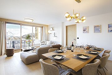 Bright, modern living/dining room with a set table, cozy sofa, and large windows looking out onto a snowy mountain landscape.