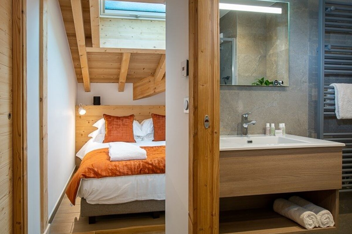 Cozy bedroom with wooden ceilings and orange bedding, next to a modern bathroom with a wooden sink vanity.