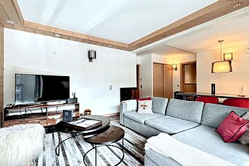 A cozy, modern living space featuring a grey sectional sofa, large TV, nested coffee tables, and a striped rug. Wooden ceiling beams add rustic charm.