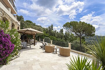 A beautiful Mediterranean villa terrace with dining and lounging areas, purple flowers, and a panoramic view of green hills under a cloudy blue sky.