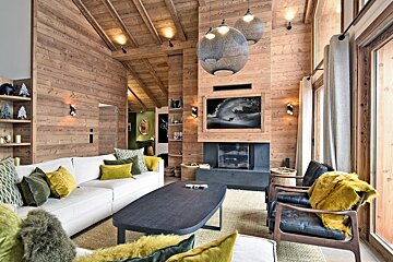A cozy, wood-paneled chalet living room featuring a white sofa, green/yellow pillows, fireplace, large windows, and woven pendant lights.
