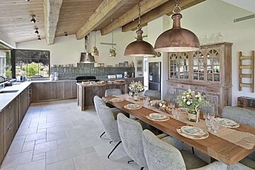 A spacious, rustic open-plan kitchen and dining room with wooden beams, copper pendant lights, and a long dining table set for guests.