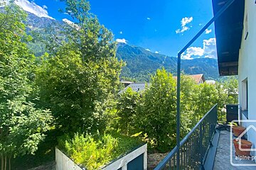 Sunny balcony view overlooking lush green trees, mountain peaks, and a bright blue sky with light clouds.