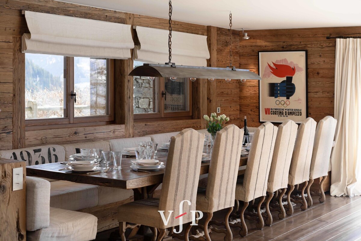 A rustic cabin dining room featuring a long wooden table, high-backed upholstered chairs, and a vintage Cortina d'Ampezzo Olympic poster.